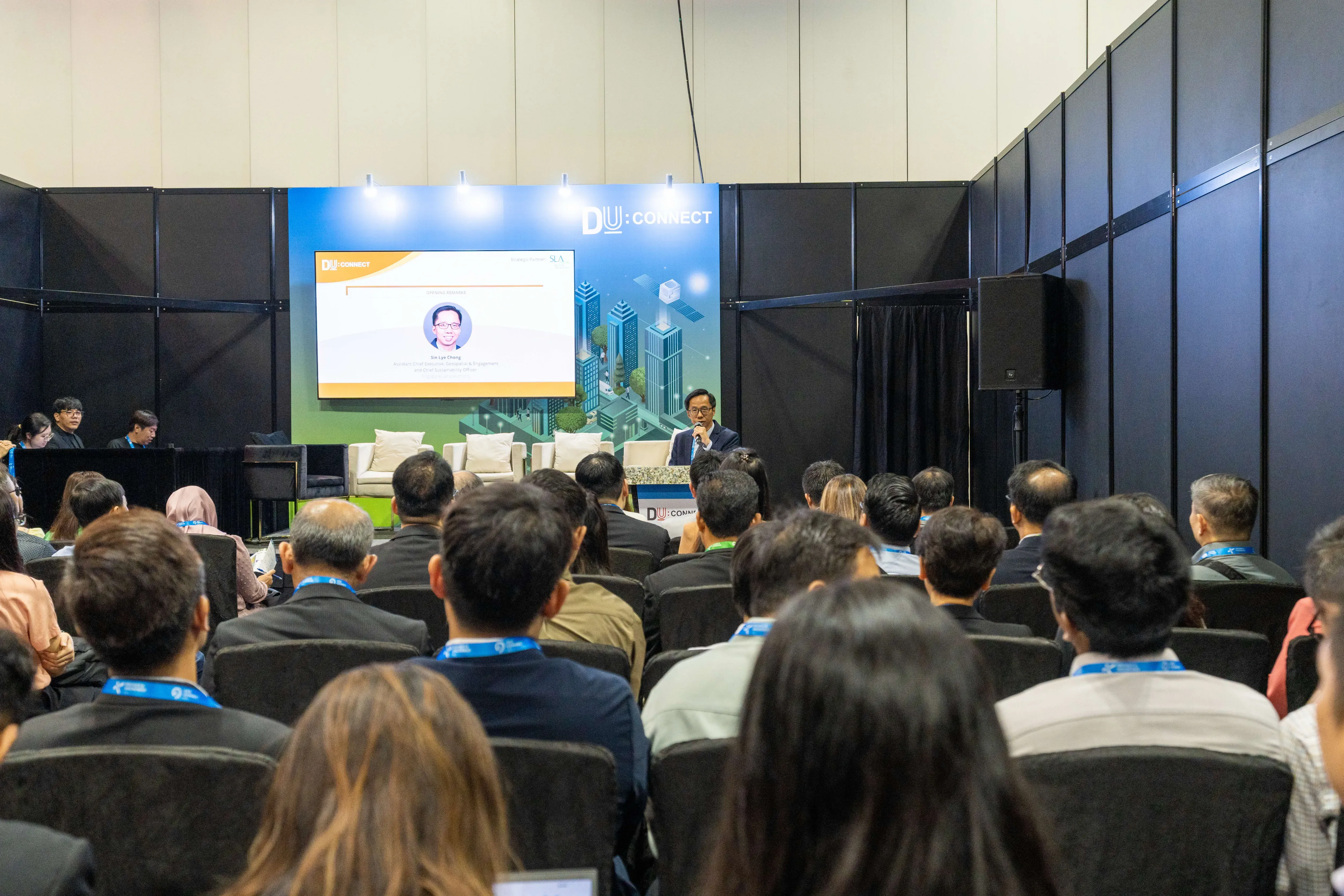 Audience seated at a conference talk facing a stage with a large DU CONNECT screen and a speaker at a podium.